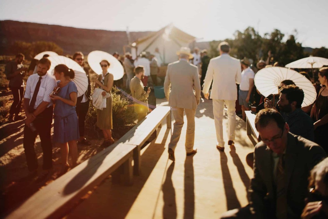 Under Canvas wedding guests wear formal attire and hold parasols outdoors on a sunny day, creating a festive atmosphere.