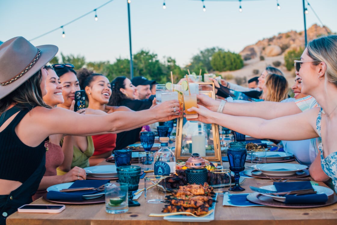 People clink glasses around a set table at an Under Canvas event with string lights above and a desert landscape behind them.