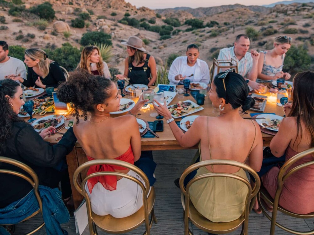 Guests at an Under Canvas event sit at a wooden table, eating and talking, surrounded by a desert landscape during sunset.
