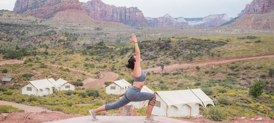 A woman performs a yoga pose on an event deck, surrounded by tents and a mountainous landscape at Under Canvas Zion.