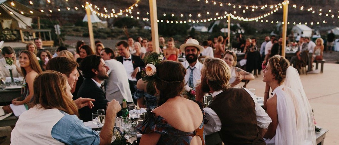 Guests at an Under Canvas wedding celebrate outdoors under string lights, surrounded by a hilly landscape.