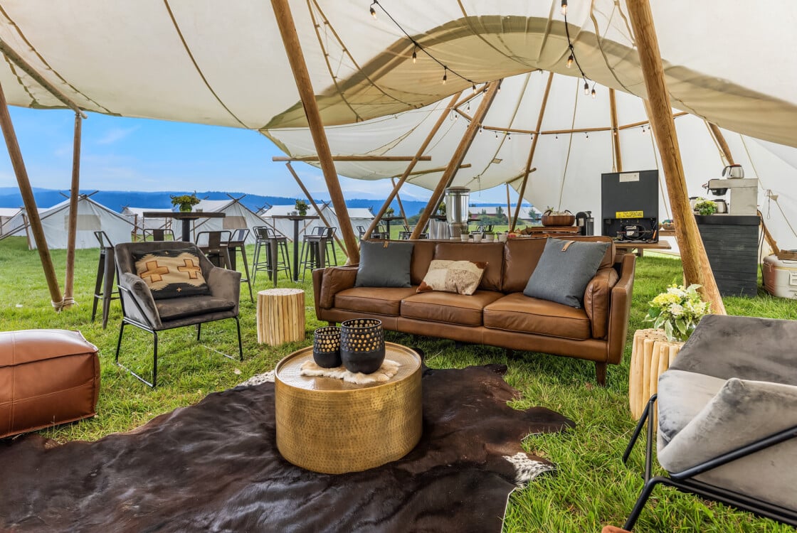 An Under Canvas event space, with a brown leather sofa under a white canvas tent, surrounded by rustic stools and a fur rug on grass.