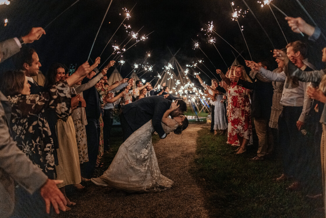 A couple kisses while surrounded by guests holding sparklers, forming an archway on a nighttime path at an Under Canvas wedding.
