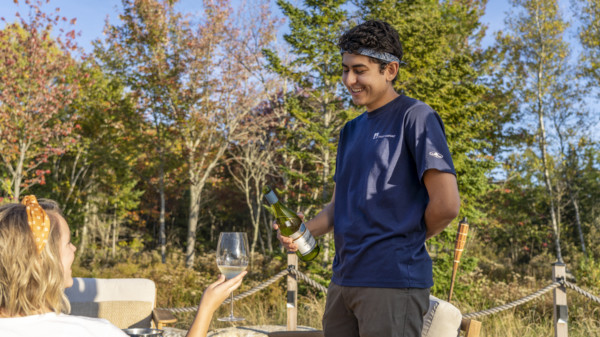 A staff member at Under Canvas serves wine, smiling at a seated individual holding a glass, surrounded by trees.