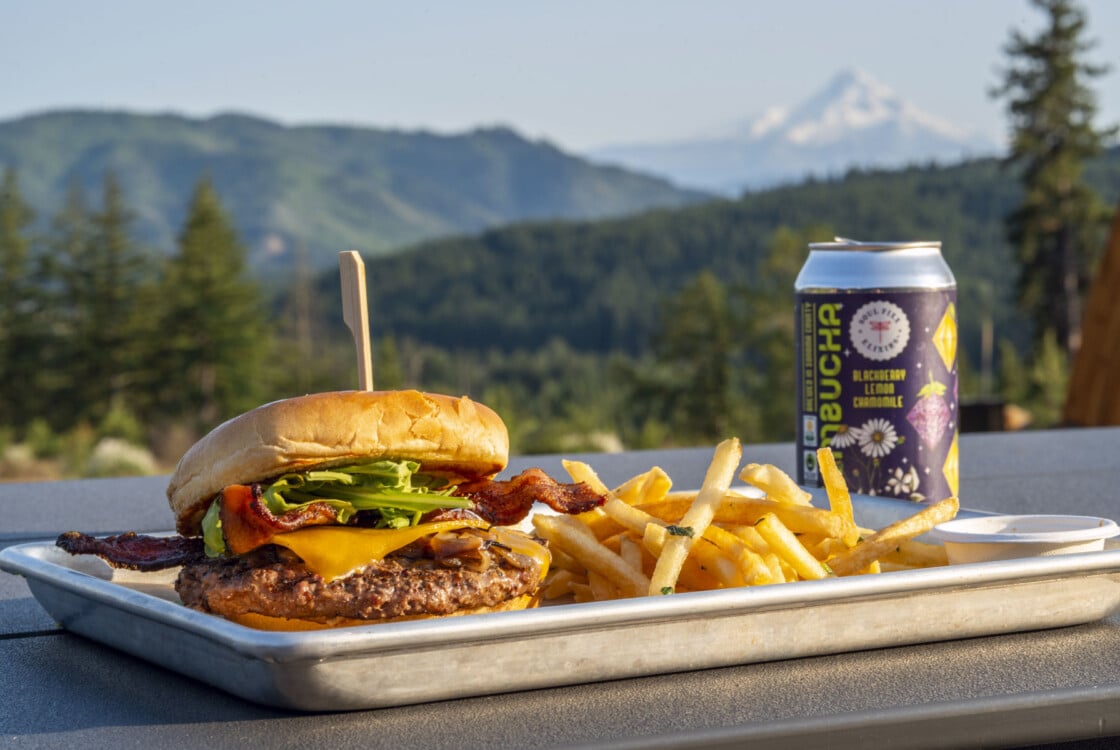 Cheeseburge with toppings, fries and a beverage sitting on a table with views of Mount Hood at Under Canvas Columbia River Gorge